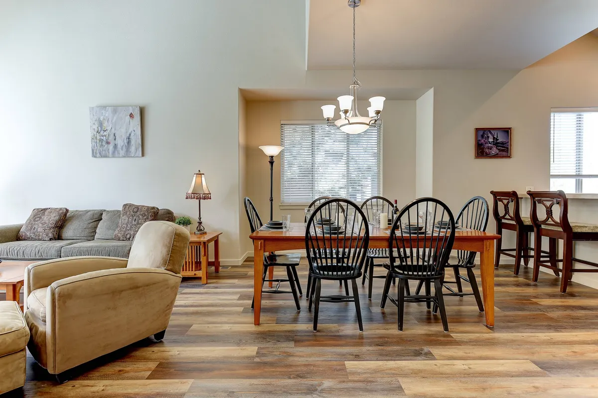 The image displays a cozy dining area with a table, sofa, chandelier, and lamp—ideal for family dinners, game nights, or casual gatherings. The ambiance is inviting and warm, suggesting comfort and togetherness.