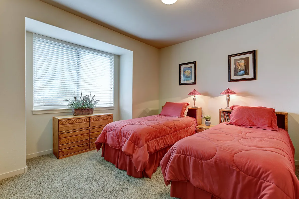 A bedroom featuring two beds with red comforters, a window with blinds, decorative lamps, framed artwork, and a wooden dresser. The room appears cozy and inviting, ideal for relaxing or conversing after a day of activities.