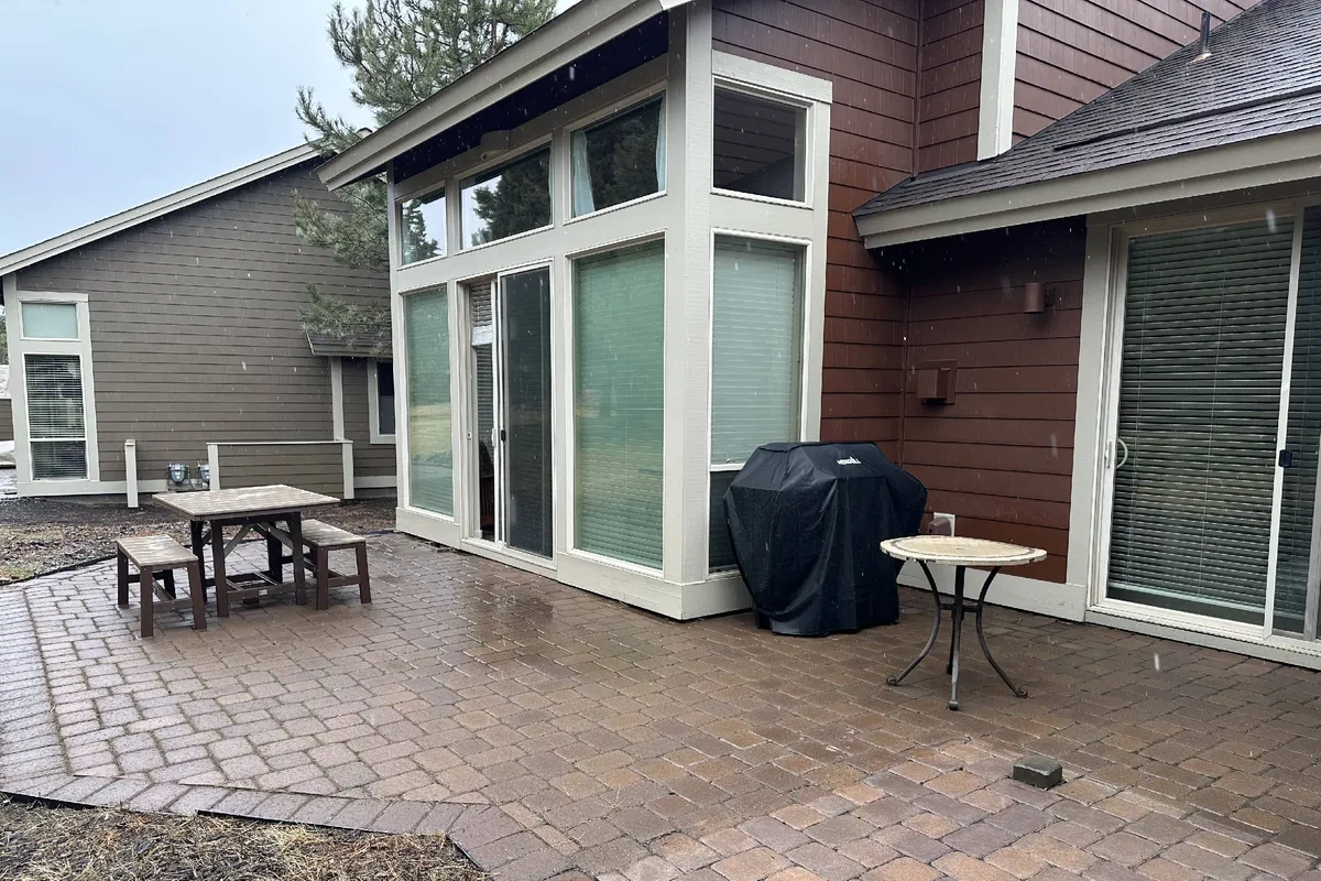 A private patio area featuring comfortable outdoor furniture under a sheltered section, with sliding glass doors opening directly from the home. The patio is set for outdoor dining, with barbecue equipment visible and natural surroundings just beyond the patio edge.