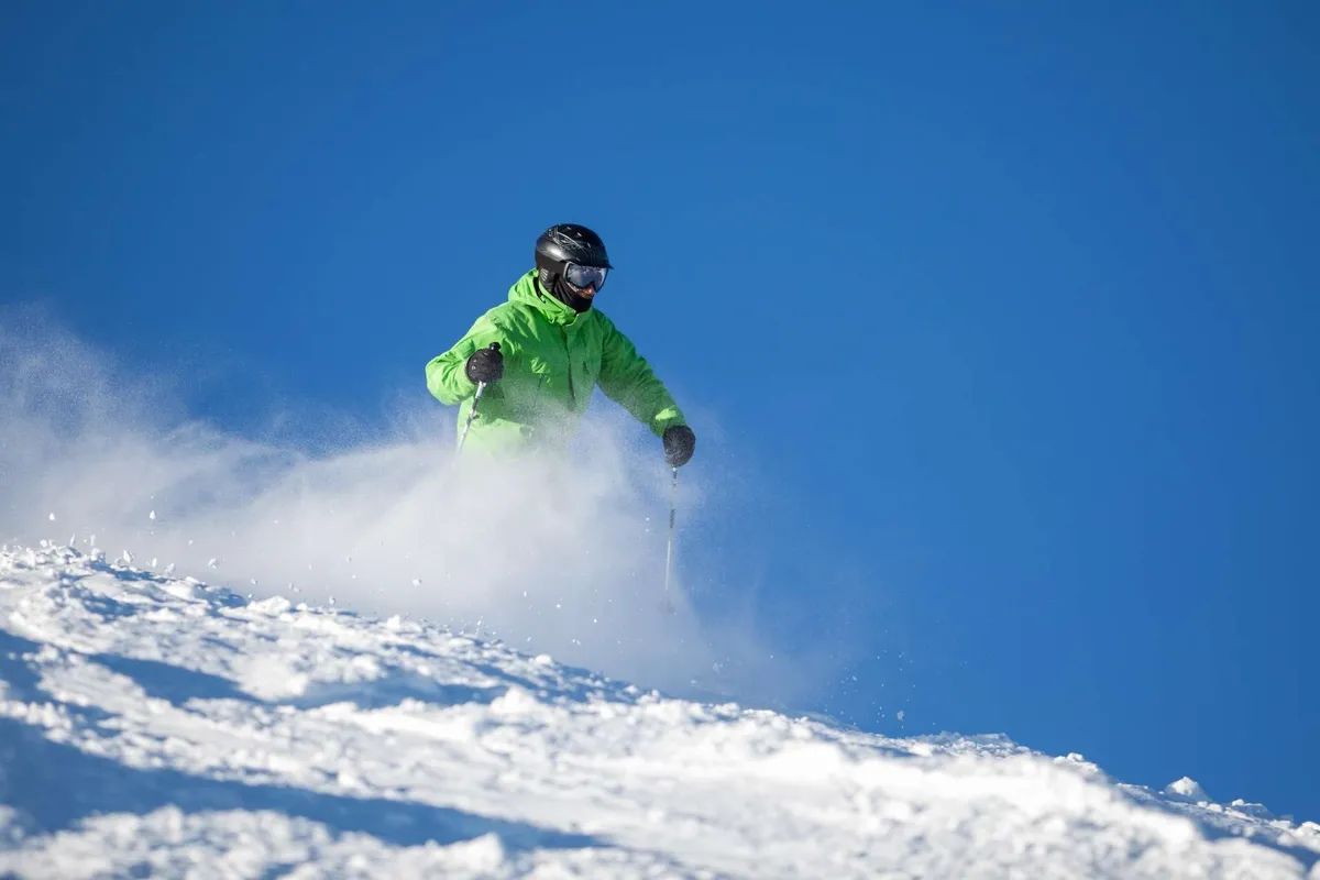 An exhilarating shot of a skier in a bright green jacket gliding down a snowy slope under a clear blue sky. The expansive mountain landscape underscores the sense of adventure and connection to nature.