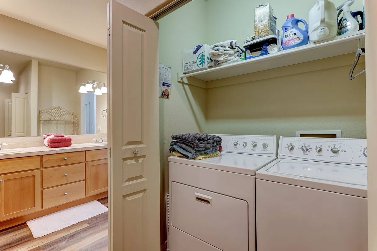 A practical and inviting view of an in-unit laundry area, complete with washing machine, dryer, and organized shelves with laundry supplies, adjacent to a bathroom vanity and mirror. The space feels efficient and welcoming, perfect for post-adventure clean-up.