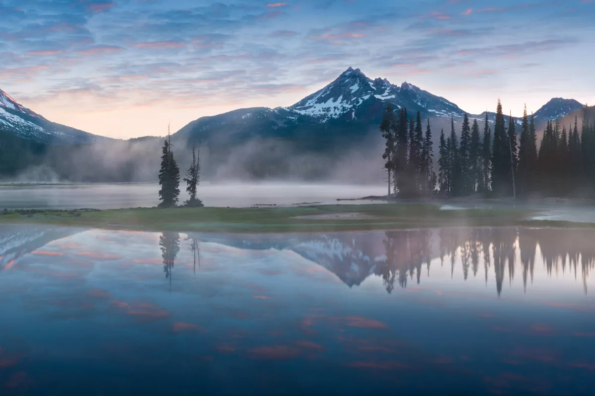A breathtaking view of snow-capped mountains reflected in a calm lake, with mist rising gently from the water and dense pine trees lining the shore. The scene is tranquil yet majestic, evoking the magic of outdoor adventures and the serenity of nature.