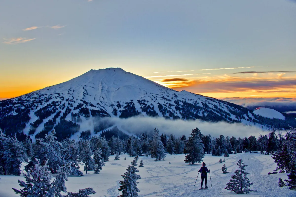 A vibrant sunset blankets the sky behind a snow-covered mountain, where a lone skier glides across the slope. The scene balances excitement and serenity, embodying the spirit of adventure and shared experiences in a stunning alpine setting.