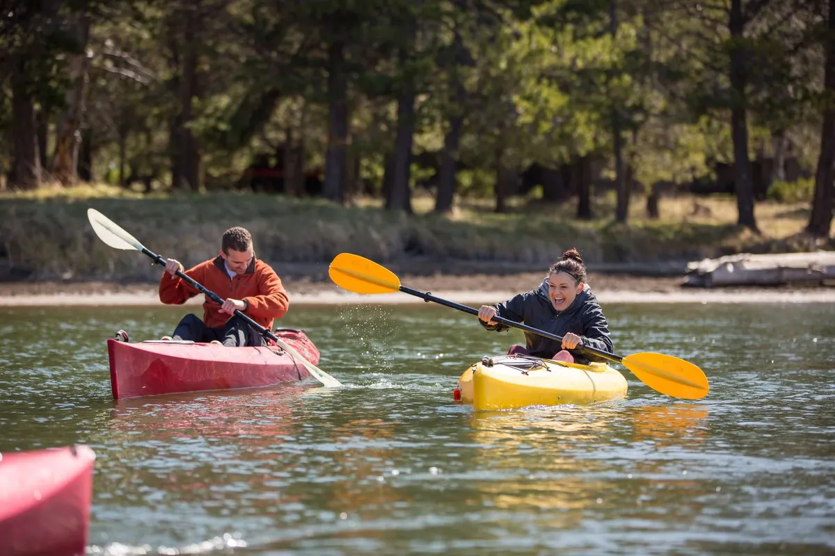 Two kayakers in brightly colored kayaks glide across a calm, reflective body of water. The scene is lively and playful, highlighting both the excitement and tranquility of water-based adventures available nearby.
