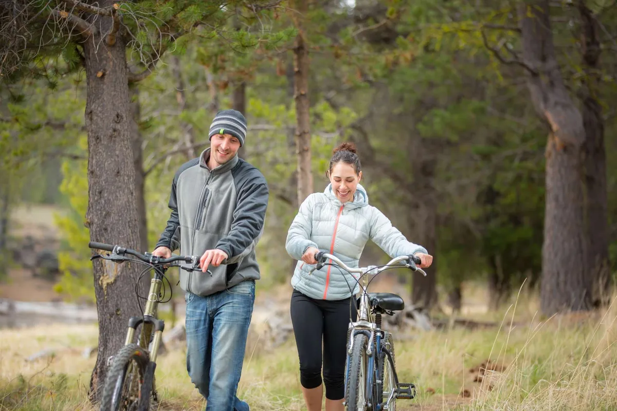 This image features a couple with bicycles enjoying a leisurely ride through a wooded area. Sunlight filters through the trees, creating a relaxed and joyful outdoor atmosphere. The scene captures the spirit of discovery and family-friendly exploration along scenic trails.